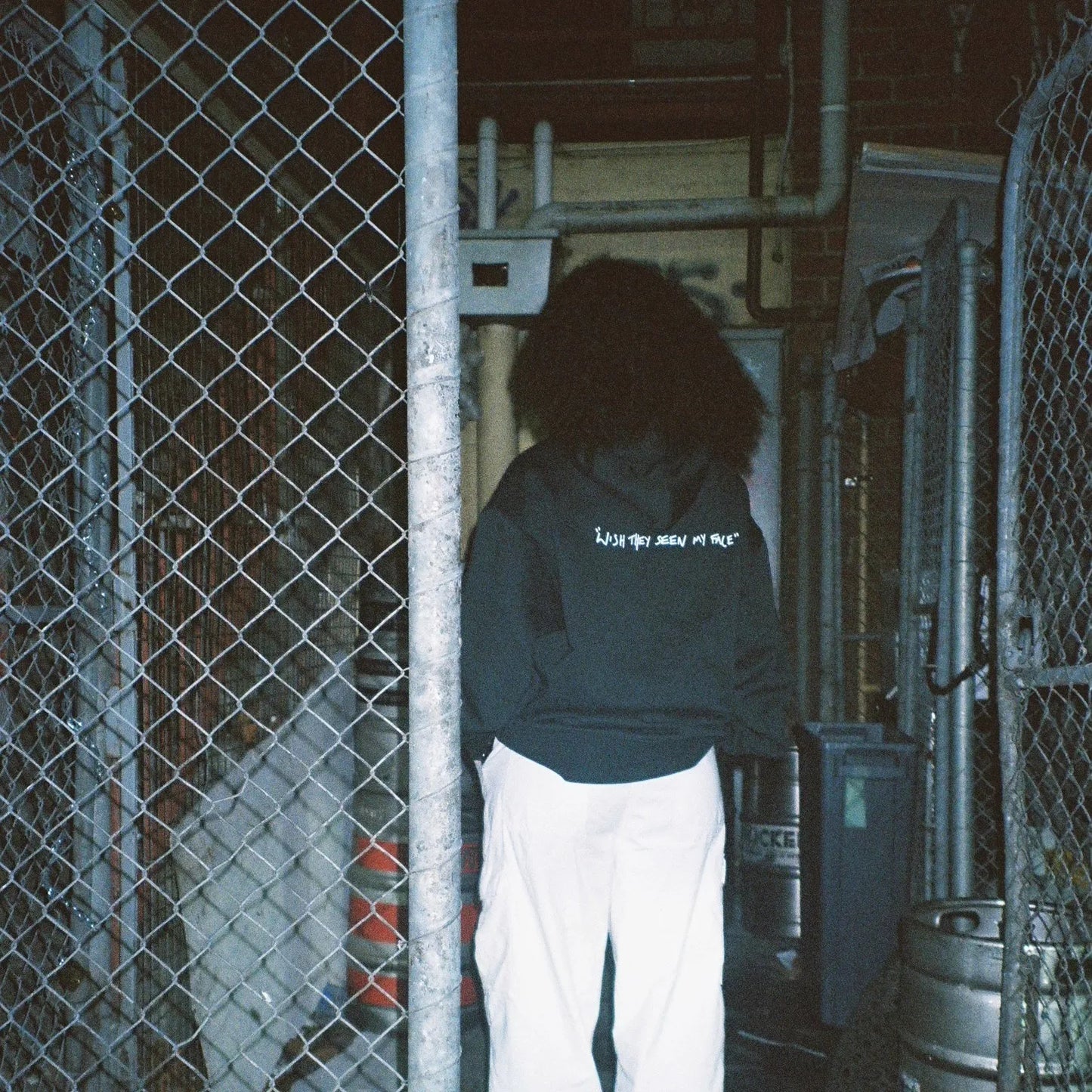 Person wearing the green Lithe hoodie with "wish they seen my face" text on the back, standing in an alleyway. Created after the popular single. Released in 2022.